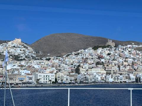 A dense hillside town of white and pastel houses rises from a deep-blue Aegean harbor under clear skies.