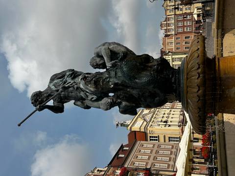 Bronze statue of Neptune atop a fountain stands in a colorful market square framed by historic townhouses.