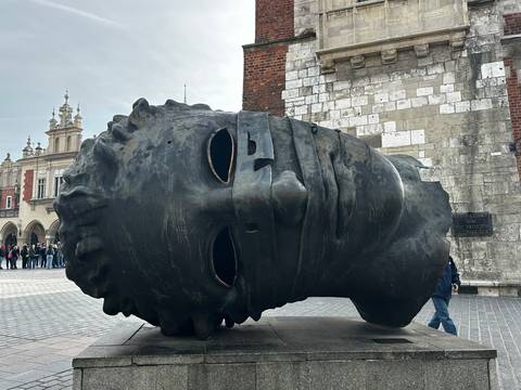 The giant bronze Eros Bendato head sculpture lies sideways in Krakow's main square as visitors walk past.