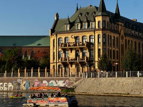 Golden evening light illuminates a riverside Neo-Gothic building above a graffiti-covered embankment.