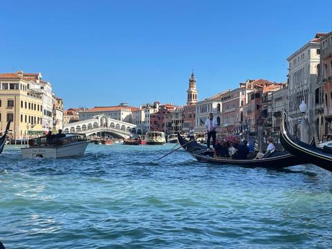 Gondolas glide along Venice’s Grand Canal with the iconic Rialto Bridge and colorful palazzi under a crisp blue sky.