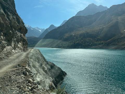Rugged cliffside track overlooks turquoise alpine lake and distant snow-capped peaks.