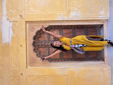Smiling woman in mustard dress raises arms in front of an ornate arched wooden doorway in a yellow palace wall.