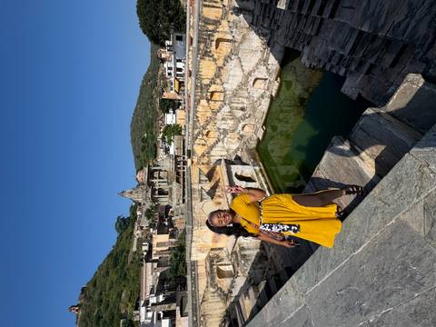 Visitor flashing a peace sign at a historic stepwell complex backed by arched pavilions and emerald water.