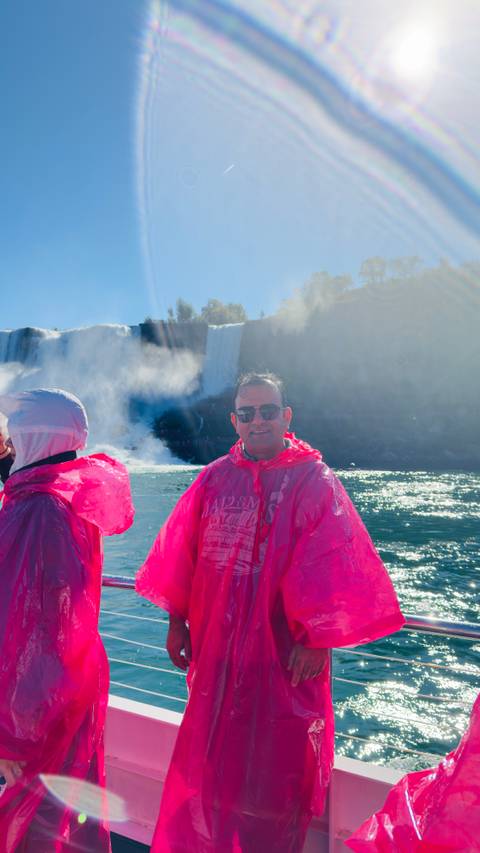 Visitors in pink ponchos stand aboard a boat near the misty torrents of Niagara Falls on a sunny day