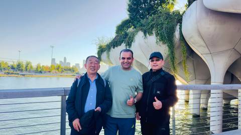 Three men smile and give thumbs-up on a riverside boardwalk with Little Island park and NYC skyline behind