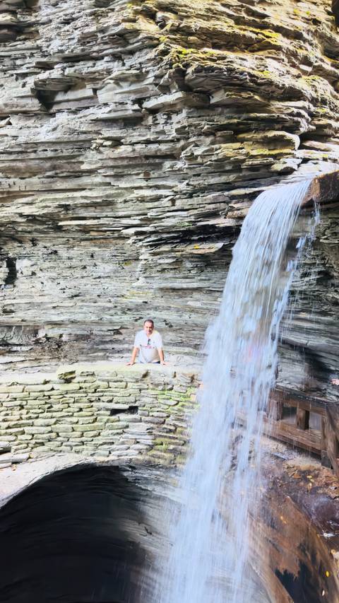 Man kneels behind a curtain of waterfall inside a rocky gorge with layered stone walls