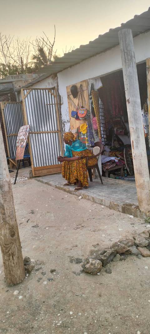 Woman in patterned dress sits on a plastic chair outside a small roadside stall made of corrugated metal