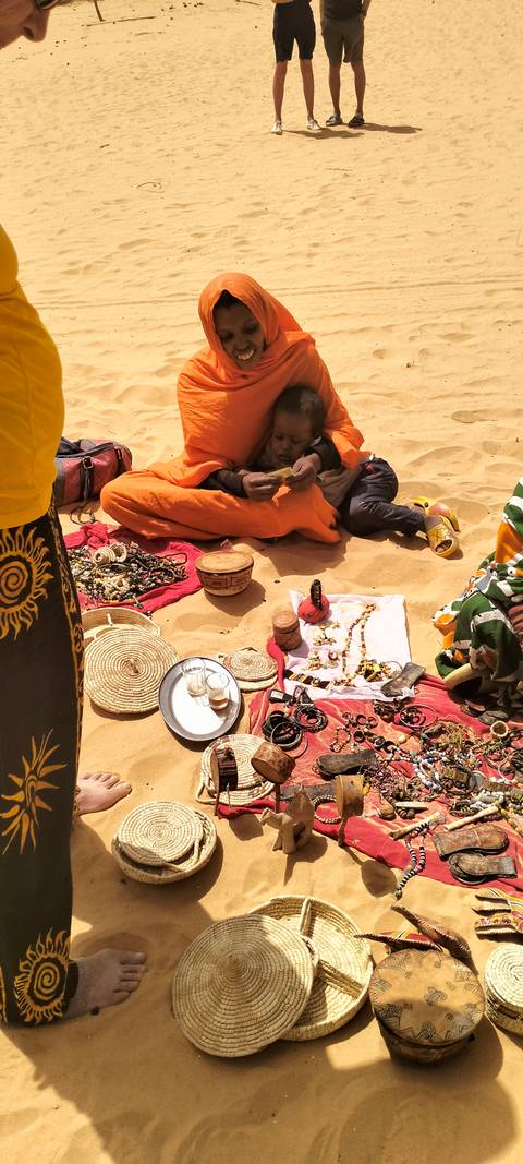 Vendor displays handmade jewelry, cups of tea and crafts on bright cloths spread over desert sand
