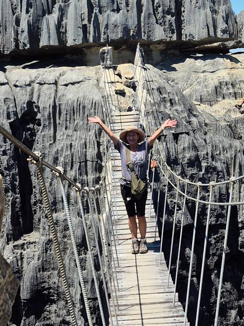 Traveller with arms raised crosses a narrow suspended wooden bridge between jagged grey limestone pinnacles.