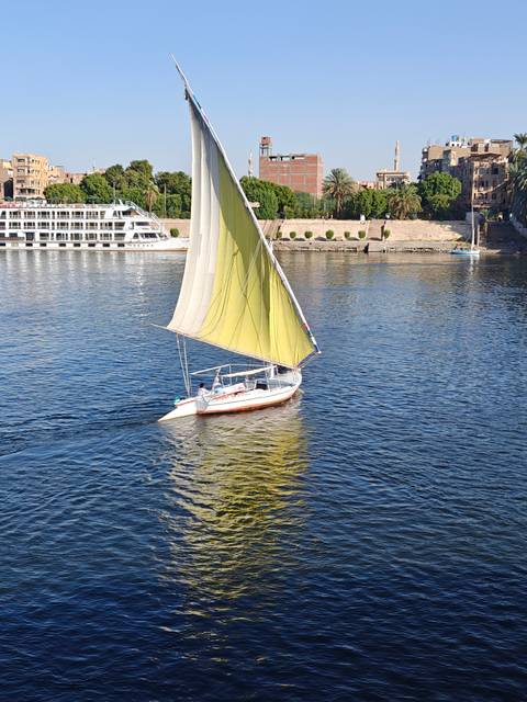 A traditional felucca with a large cream-colored sail glides over the calm blue Nile River.