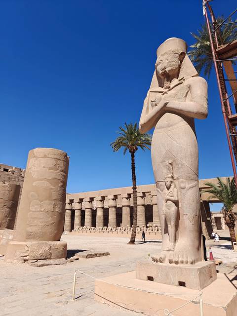 A tall pharaoh statue and palm tree rise against a vivid blue sky amid Karnak Temple columns.