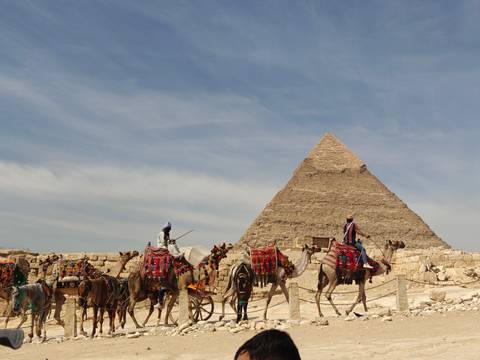 Camel riders in colorful blankets pass in front of the Great Pyramid under a lightly clouded sky.