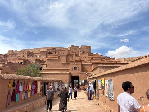 Adobe-brick ksar of Aït Benhaddou rises above market stalls and tourists under blue sky.