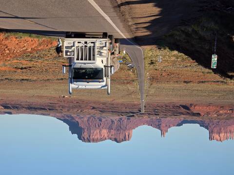 A long straight highway leads toward Monument Valley’s red buttes, with a white semi-truck in the foreground.