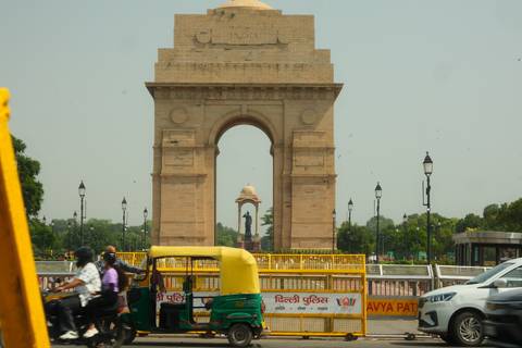 India Gate monument framed by busy city traffic and hazy sky, with auto-rickshaw in foreground.