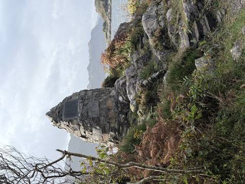 Stone cairn memorial on rocky terrain surrounded by ferns and shrubs with distant mountains.