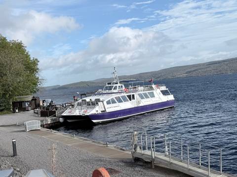 Passenger tour boat moored at a pier on a deep blue loch under partly cloudy skies.
