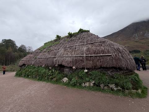 Traditional thatched blackhouse structure with visitors standing nearby under overcast skies and misty mountains.