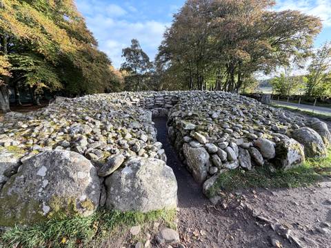 Circular prehistoric stone burial cairn constructed of lichen-covered rocks surrounded by autumn trees.