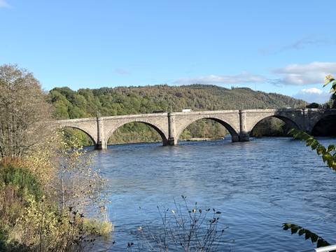 Historic stone arch bridge spanning a wide river bordered by forested hills under clear blue skies.