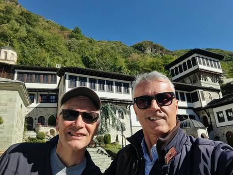Two travellers taking a selfie in front of an ornate Macedonian monastery complex set against forested hills.
