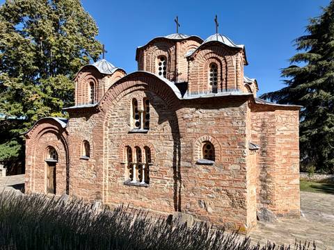 Small medieval brick church with curved rooflines in a shaded garden setting.