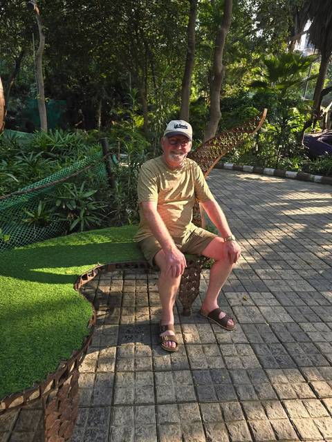 Older man sitting on a leaf-shaped bench in a sunny garden path.