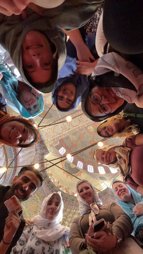 Creative upward selfie of friends in headscarves forming a circle beneath the ornate tiled ceiling of a mosque.