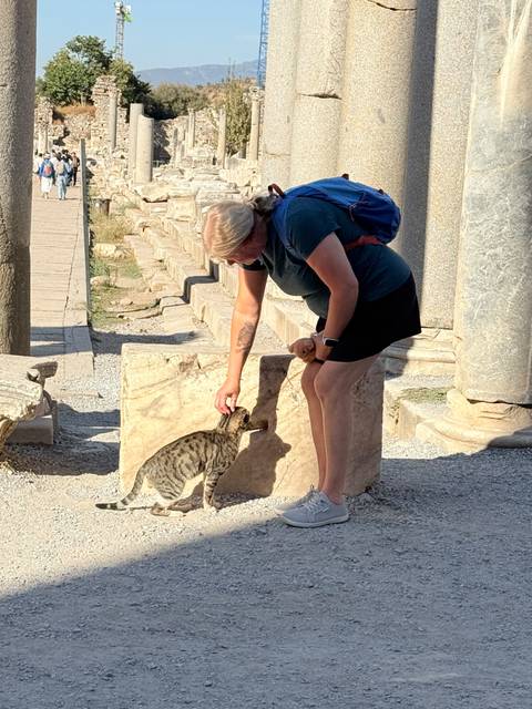 Traveler bends to pet a striped cat among marble ruins and fallen columns on a sunny day.