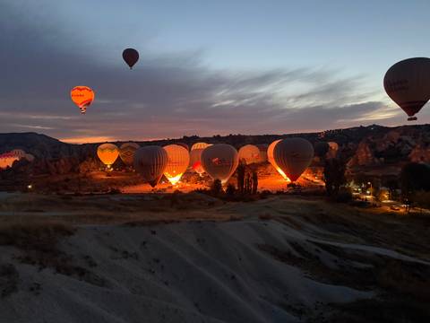 Pre-dawn scene of tethered hot air balloons glowing orange as their burners ignite against a purple sky.