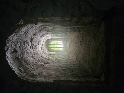Rough stone tunnel interior leads to barred window with greenery beyond.