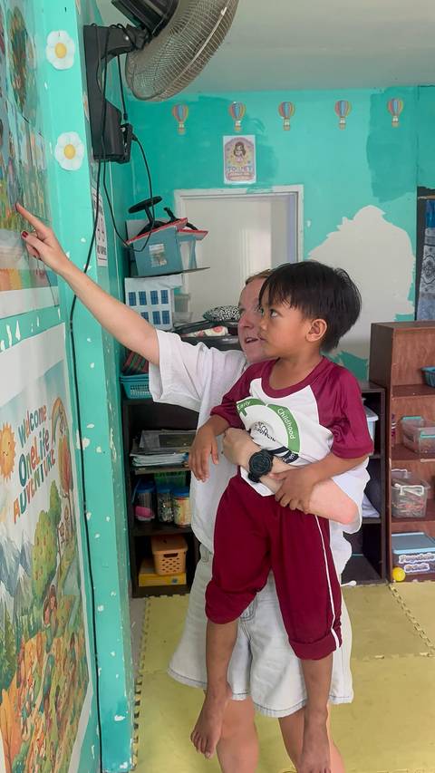 A volunteer teacher helps a young child point at colorful posters inside a classroom.