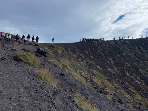Hikers walking along the rim of a volcanic crater with dark lava rocks and sparse vegetation.