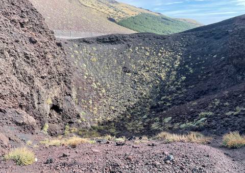 Deep volcanic crater with dark lava rocks and sparse vegetation in bright light.