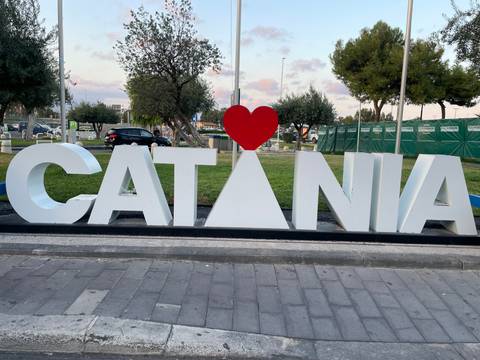 Large white and red love-themed CATANIA sign near a city street with trees behind.