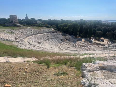 Wide view of ancient stone Greek theatre carved into hillside surrounded by greenery.
