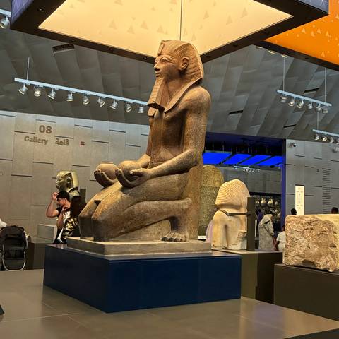 Indoor gallery scene featuring a large kneeling ancient Egyptian statue with visitors observing exhibits.