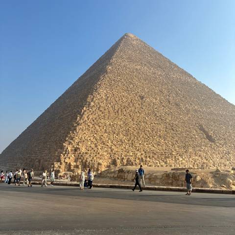 Wide view of the Great Pyramid with tourists walking around its base under bright blue sky.
