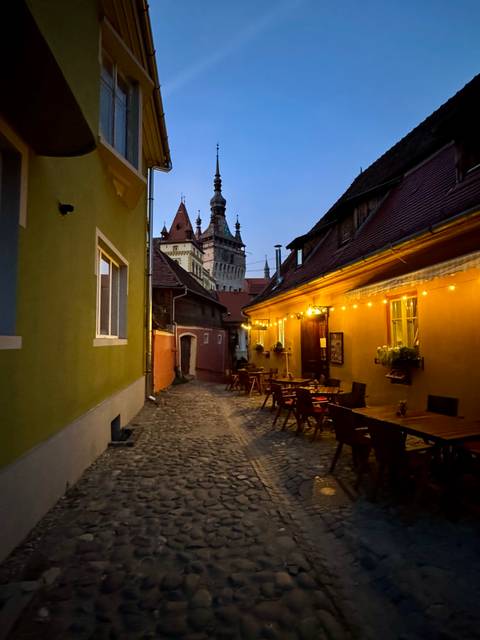 Cobblestone alleyway in Sighisoara illuminated by string lights with outdoor tables at dusk.