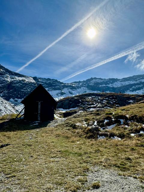 Small wooden hut on snowy alpine ridge with jagged peaks and vapor trails overhead.