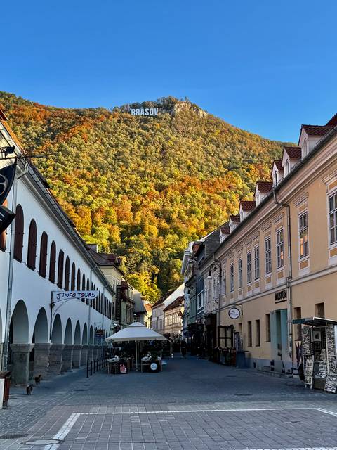 Street of pastel buildings opening to forested hillside ablaze with autumn colors in Brasov.