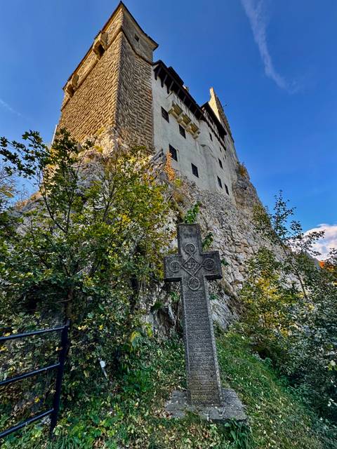 Upward view of stone walls of Bran Castle with ornate cross monument among foliage.