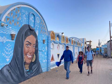Tourists walk past vibrant blue Nubian village walls adorned with large mural art and geometric decorations.