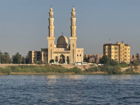A mosque with twin minarets and a central dome stands on the Nile riverbank viewed from across the water.