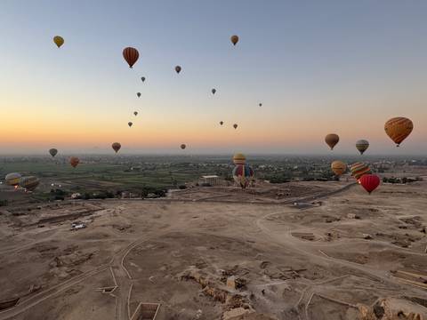 Dozens of colorful hot-air balloons rise over the ruins of Luxor at dawn with orange sky above farmland.