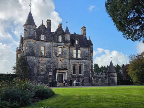 Victorian-era manor house with turrets and slate roof against a bright sky, visitors at entrance.