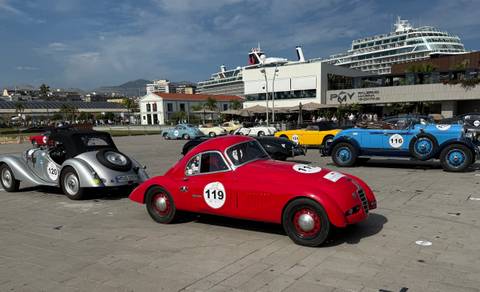 Classic sports cars displayed on a waterfront plaza with a docked cruise ship towering behind