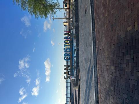 Colourful SAN CRISTOBAL sign on a waterfront promenade under a blue sky with scattered clouds.