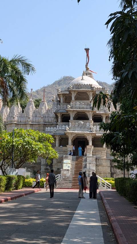 Intricately carved white marble Jain temple complex set against a hillside with visitors at the entrance.